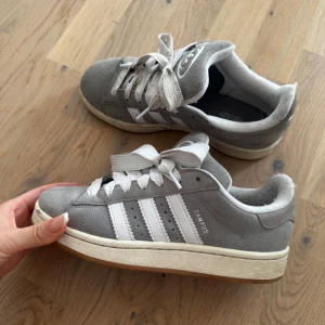 Grå Adidas Campus sneakers i mocka - Snygga grå Adidas Campus sneakers med klassiska vita ränder och logga på hälen. Skorna har en låg profil, rund tå och platt sula. Ovandelen är i mjuk mocka och de har vita snören. Perfekta för en avslappnad streetstyle-look.