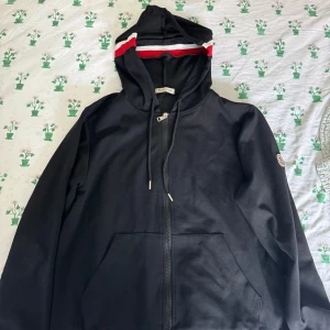 Svart Moncler hoodie med dragkedja - Svart hoodie från Moncler med hel dragkedja, huva och snörning. Ribbade muddar med röd och vit rand, samt Moncler-logga på ärmen. Tillverkad i mjuk bomull, perfekt för en chill och stilren look.