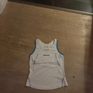 Träningslinne - Säljer en vit linnetopp från Babolat med blå detaljer och sportig design. Toppen har ärmlös modell och rund halsringning, perfekt för dig som gillar att spela tennis eller träna. Materialet är lätt och andas, vilket gör den skön att ha på sig under aktivitet.
