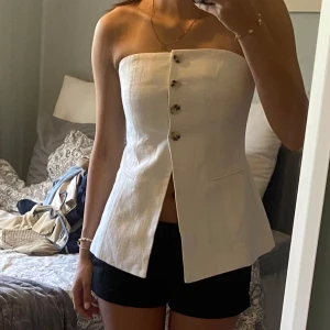 Vit bandeau tubtopp med knappar - Säljer en stilren vit bandeau tubtopp med snygg knappdetalj framtill och slits nedtill. Toppen är axelbandslös och har en figurnära passform som ger en clean och modern look. 