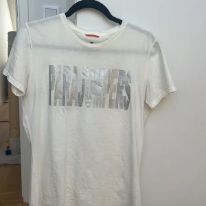 Vit t-shirt från Parajumpers - Vit t-shirt från Parajumpers med stort, silvrigt logotryck över bröstet. Klassisk rund halsringning och korta ärmar. Skön och lätt i bomull, perfekt till jeans eller shorts. Snygg och enkel stil med statement-tryck. Är 164 och passar mig 