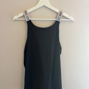 Svart Nike linnetopp med logoband - Sportig svart linnetopp från Nike med breda vita axelband med röd Nike-logga och blå ränder. Toppen har en blå Nike Swoosh framtill och är tillverkad i Dri-FIT-material som andas. Perfekt för träning eller när du vill ha en clean och cool look.