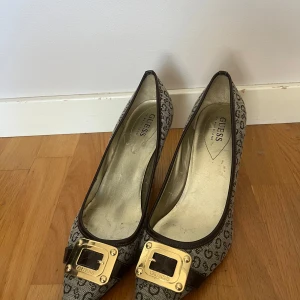 Guess klackar med monogram och gulddetalj - Snygga klackar från Guess med beige och brunt monogrammönster, spetsig tå och låg klack. Skorna har en stor guldfärgad spänne framtill med Guess-logga och bruna lackdetaljer. Tillverkade i textil med syntetdetaljer. skriv för fler bilder❤️