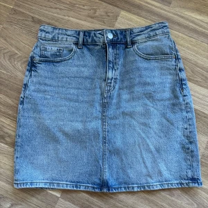 Blå jeanskjol från VILA, strl 38 - Klassisk blå jeanskjol från VILA i storlek 38. Modellen är kort med fem fickor, bälteshällor och knapp framtill. Perfekt till sneakers eller boots för en chill look.