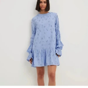 Ljusblå volangklänning med blommor - Använd fåtal gånger! Supersöt ljusblå kortklänning med volang nertill och långa ärmar med volangdetaljer. Klänningen har små blommiga broderier över hela tyget och en avslappnad passform. Perfekt för vår och sommar!