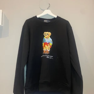 Svart sweatshirt Polo Bear Ralph Lauren - Svart sweatshirt från Ralph Lauren med ikoniskt Polo Bear-tryck framtill. Tröjan har rund halsringning och långa ärmar. Perfekt för dig som gillar streetstyle och vill ha något som sticker ut med färgglad print.