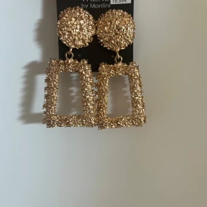 Guldiga statementörhängen Montini - Stora statementörhängen från Montini i guldig färg. Örhängena har en rund, texturerad topp och en rektangulär, hängande del. Perfekta för att sticka ut och ge din look extra edge.