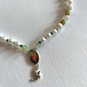Pärlhalsband med duva - Unikt halsband med vita pärlor, små gröna och blå pärlor samt detaljerade rosformade pärlor. I mitten hänger en religiös medaljong med motiv och en liten silverfärgad duva. Perfekt statement-piece för dig som gillar personliga smycken.