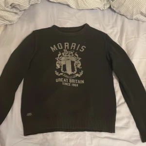 Mörkblå tröja från Morris - Säljer en snygg mörkblå tröja från Morris med tryck framtill och ett broderat brittiskt flaggmärke på ärmen. Tröjan har ribbade muddar vid ärmslut och nederkant, vilket ger en stilren look. Perfekt för dig som gillar klassisk design med en twist!
