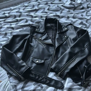 Svart skinnjacka från Zara, S - Snygg svart skinnjacka från Zara med dragkedjor, fickor och bälte i midjan. Jackan har klassisk krage med nitar och detaljerade sömmar.