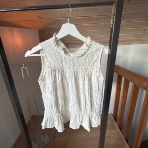 Vit ärmlös blus med spetsdetaljer - Supersöt vit blus med volangkant nertill och broderade spetsdetaljer upptill. Den är ärmlös och har en lätt rynkad struktur i tyget som ger en romantisk vibe. Perfekt för dig som gillar en feminin och luftig stil. Från Zara