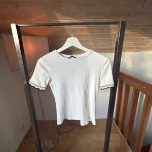 Vit topp med spetsdetalj från Zara - Söt vit topp från Zara med korta ärmar och rund halsringning. Ärmsluten har fina spetsdetaljer i svart och guld som ger en unik touch. Perfekt för dig som gillar stilrena plagg med en twist.