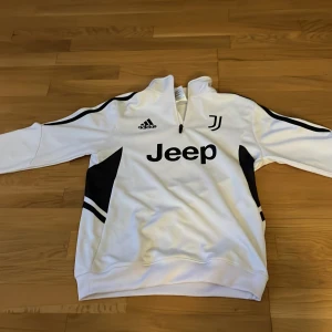 Vit Juventus half zip tröja Adidas - Säljer en vit långärmad Juventus-tröja från Adidas med svart Jeep-logga och klubbmärke på bröstet. Tröjan har svartvita detaljer på sidorna och ärmarna samt en praktisk half zip vid halsen. Perfekt för fotbollsträning eller chill.