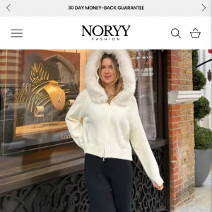 Vit stickad jacka med fuskpälsluva - Mysig vit stickad jacka med dragkedja och stor luva kantad av fluffig fuskpäls. Jackan har ribbade muddar och en croppad passform som ger en trendig look. Perfekt för kyliga dagar när du vill vara både varm och snygg.