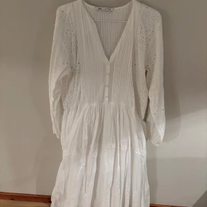 Vit långklänning med broderi från Zara - Superfin vit långklänning från Zara med broderade detaljer och hålmönster. Klänningen har v-ringning, knappar framtill och långa ärmar. Tillverkad i bomull, perfekt för sommarens alla tillfällen. Luftig och romantisk stil med volang och spetsdetaljer.