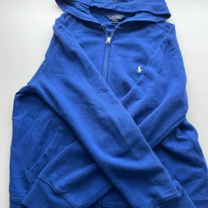Blå hoodie från Polo Ralph Lauren - Blå hoodie från Polo Ralph Lauren med dragkedja framtill och klassisk gul logga broderad på bröstet. Tröjan har huva, långa ärmar och känguruficka. Perfekt för en chill och sportig stil.