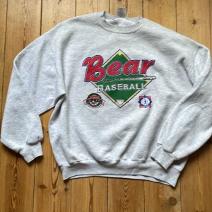 Grå Bear Baseball sweatshirt Jerzees XL - Snygg grå sweatshirt från Jerzees i storlek XL med stort Bear Baseball-tryck framtill i rött, grönt och blått. Tröjan har rund hals, muddar vid ärmslut och nederkant. Materialet är en mix av bomull och polyester, perfekt för dig som gillar sportig och avslappnad stil.