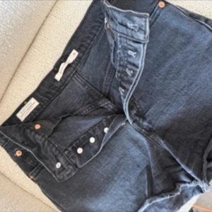 Svarta jeansshorts från Levi's - Snygga svarta jeansshorts från Levi's med hög midja och klassisk knappgylf. Shortsen har råa kanter nertill för en cool, avslappnad look. Perfekta för sommardagar och festivaler.