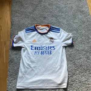 Real Madrid Adidas fotbollströja vit - Säljer en officiell Real Madrid fotbollströja från Adidas. Tröjan är vit med blå och orange detaljer på axlar, krage och ärmslut. Klassisk passform med korta ärmar och klubbmärke på bröstet. Materialet är lätt och andas, perfekt för match eller träning.