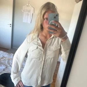 Beige skjorta från Aware med fickor - Stilren beige skjorta från Aware med två bröstfickor och klassisk krage. Skjortan har långa ärmar med manschett och knappar hela vägen framtill. Materialet känns lätt och luftigt, perfekt till jeans eller kjol. Enkel och clean look.