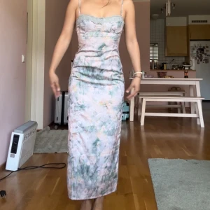 Pastellfärgad långklänning med spets - Säljer en långklänning i pastelliga toner med blommigt mönster och tunna axelband. Klänningen har spetsdetalj upptill, figurnära passform och slits bak. Materialet känns mjukt och svalt, perfekt för sommarens alla tillfällen.