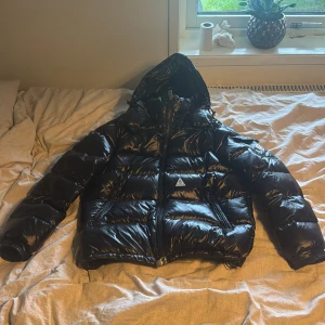 Svart Moncler pufferjacka med huva - Säljer en svart, glansig pufferjacka från Moncler med huva och dragkedja. Jackan har klassisk Moncler-logga på ärmen och coolt serietidningsfoder på insidan. Perfekt för kalla dagar och riktigt snygg streetstil.