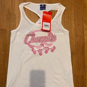 Vit linnetopp från Champion - Snygg vit linnetopp från Champion med rosa tryck framtill som visar rullskridskor och logga. Toppen har brottarrygg med twistad detalj bak och ärmlös design. Perfekt för en sportig och avslappnad look.