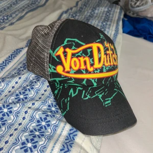 Vintage Von Dutch keps med grafiskt tryck - Säljer en svart och grå vintage Von Dutch keps med färgstarkt gult och rött logotryck framtill samt grönt grafiskt mönster. Klassisk trucker-modell med mesh på sidorna och justerbar baksida. Tillverkad i 100% polyester och bomull.