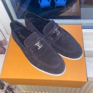Marinblå mockaloafers från Louis Vuitton - Snygga marinblå loafers från Louis Vuitton i mjuk mocka med silverfärgad LV-detalj framtill. Skorna har en klassisk rund tå och vit sula, perfekt för dig som gillar stilrena och exklusiva skor. Kommer med originalkartong och kvitto.