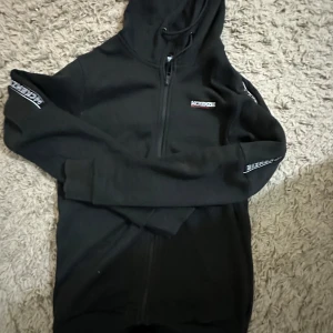 Svart hoodie från McKenzie med dragkedja - Svart hoodie från McKenzie med hel dragkedja och huva. Snygga vita McKenzie-loggor längs båda ärmarna och på bröstet. Tillverkad i mjuk bomullsmix, perfekt för chill eller streetwear. Långärmad och med ribbade muddar för extra komfort.