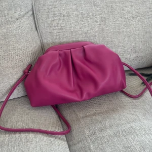 Rosa axelväska i skinn med veck - Snygg axelväska i stark rosa färg med mjukt, veckat skinn och lång axelrem. Väskan har ett rymligt innerfack med vit blommig textil och dragkedja. Perfekt för dig som vill ha en färgklick till outfiten och gillar stilren design med twist.