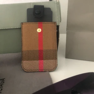 Rutig plånbok från Burberry - Snygg liten plånbok från Burberry i klassiskt rutigt mönster med bruna och röda toner. Praktisk knappstängning i svart och stilren design som passar perfekt för kort eller småsaker. Ikonisk accessoar med lyxig känsla.