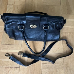 Svart skinnhandväska med axelrem - Stilren svart handväska i skinn med både handtag och avtagbar axelrem. Väskan har ett klassiskt rektangulärt snitt, snygga metalldetaljer i silverfärg och praktiskt vridlås framtill. Invändigt finns dragkedjefack och flera småfack för organisering.