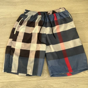 Burberry shorts - Snygga shorts med rutigt mönster i blått, svart, beige och rött. De har elastisk midja med svart snörning och sidofickor. Perfekta för sommaren och har en loose passform. Materialet känns lätt och syntetiskt, vilket gör dem sköna att bära varma dagar.