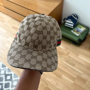 Gucci keps med GG-mönster och ränder - Snygg keps från Gucci i beige med klassiskt GG-monogram över hela kepsen. På sidorna finns de ikoniska gröna och röda ränderna. Justerbar rem baktill i mörkbrunt skinn. Perfekt accessoar för att lyfta din outfit.