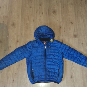 Blå dunjacka från Parajumpers - Snygg blå dunjacka från Parajumpers med huva och coolt märke på ärmen. Jackan är superlätt, har dragkedja framtill och två fickor med dragkedja. Perfekt för dig som vill ha en stilren och sportig look under kyliga dagar.