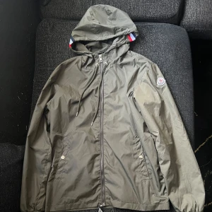Grå vindjacka från Moncler med huva - Snygg grå vindjacka från Moncler med huva och dragkedja framtill. Jackan har två fickor med tryckknappar, elastiska muddar och coola Moncler-detaljer på ärmen och insidan. Perfekt för dig som vill ha en stilren och sportig look.