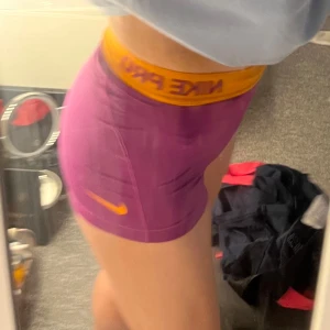 Nike Pro lila träningstights-shorts - Säljer ett par tighta lila Nike Pro shorts med orange resår och logga. Perfekta för träning eller gym, sitter högt i midjan och är gjorda i ett stretchigt syntetmaterial som andas. Snygg sportig look och riktigt bekväma att röra sig i.
