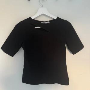 Svart topp med cut out-detalj - Snygg svart topp från Kappahl med unik cut out-detalj vid halsen. Modellen har korta ärmar och en tight passform som ger en modern och stilren look. Perfekt för dig som gillar minimalistisk stil med en twist.
