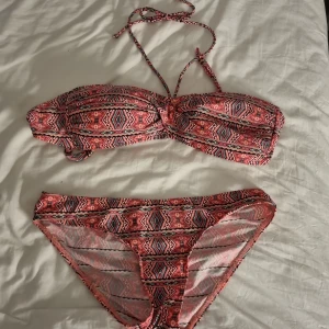 Mönstrad bikini från Andy by Frank Dandy - Snygg bikini med bandeau-topp och klassiska bikinitrosor i rosa, svart och vit med grafiskt mönster. Toppen har knytband i nacken och vadderade kupor. Materialet är stretchigt och består av polyamid, elastan och polyester. Perfekt för strandhäng och poolparty!