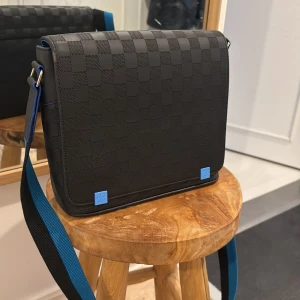 Louis Vuitton rutig axelväska svart - Snygg axelväska från Louis Vuitton i svart skinn med klassiskt rutigt Damier-mönster. Väskan har blå detaljer och en bred axelrem i svart och blått. Perfekt storlek för vardagens essentials och en stilren look.