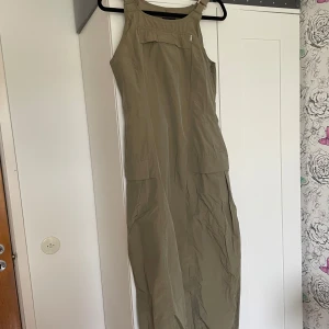Smutsgrön långklänning  - Långklänning från X-CEPT med raka linjer och breda axelband med spännen. Klänningen har fickor framtill och på sidan samt en dragkedja i sidan. Tunt material med 90s känsla.