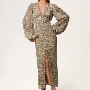Zebramönstrad långklänning med v-ringning - Snygg långklänning med zebramönster i beige och grå toner. Klänningen har djup v-ringning, långa puffärmar och knappar hela vägen framtill samt en slits. Perfekt för dig som vill sticka ut med en trendig look.