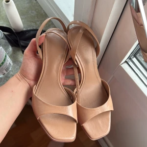Beige sandaler med smala remmar och klack - Snygga beige sandaler med smala remmar och öppen tå. Skorna har fyrkantig tå, hög klack och justerbart spänne vid ankeln. Tillverkade i syntetmaterial med stilren och minimalistisk design, perfekta för dig som gillar en clean look.