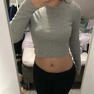 Grå croppad långärmad tröja från AYA - Säljer en grå croppad långärmad tröja från AYA med ribbad struktur. Tröjan har en tight passform och rund halsringning, perfekt till jeans eller kjol. Broderad logga bak i nacken för en extra detalj. Superenkel att styla och riktigt skön på❤️