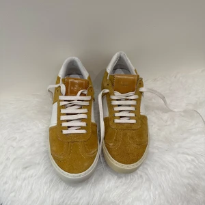 Valentino Garavani sneakers mocka/skin - Snygga sneakers från Valentino Garavani i senapsgul mocka och vitt skinn. Klassisk låg modell med vita snören, rund tå och coola nitar på sulan. Sulan har svart mönster undertill. Perfekta för dig som vill sticka ut med stilrena detaljer.