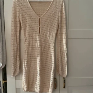 Beige stickad strandklänning med v-ringning   - Supersöt beige stickad  kort strand klänning  med v-ringning och lång ärm. Klänningen har ett hålmönster och två små öppningar framtill som ger en unik touch. 