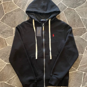 Svart hoodie från Polo Ralph Lauren - Svart hoodie från Polo Ralph Lauren med dragkedja och vit snörning i huvan. Klassisk röd broderad logga på bröstet och känguruficka framtill. Tillverkad i mjuk bomullsmix, perfekt för en chill och stilren look. 