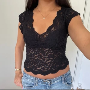 Svart spetsblus med v-ringning - Supersnygg svart blus i spets med v-ringad hals och korta ärmar. Transparent material med vågig kant nertill och vid ärmarna. Perfekt att styla med jeans eller kjol för en trendig look.