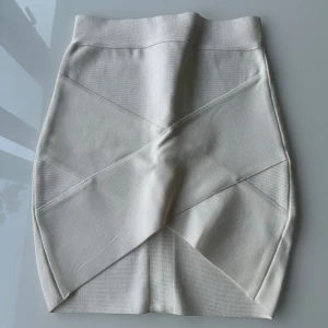 Vit bandage kjol - Snygg vit kortkjol med tight passform och coola sömdetaljer framtill. Kjolen har en dold dragkedja bak och är gjord i ett stretchigt material som sitter perfekt. Perfekt för dig som gillar stilrena och trendiga plagg. Liten i storlek!